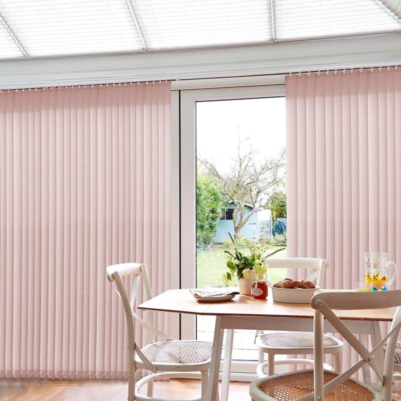 Pale pink vertical blinds covering a sliding glass door, slightly parted at center to reveal a garden; filtering soft daylight into a dining area with wooden table, chairs, and flowers.