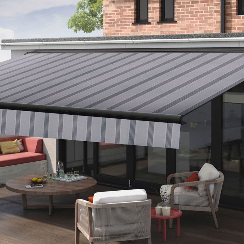 A diagonal-striped retractable fabric awning with a striped valance is fully extended from the house eave, shading a glass-walled patio furnished with low table, sofa and armchairs in daylight.