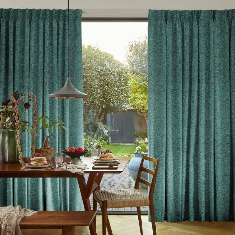 Teal textured curtains, hung to the sides and pulled open, framing an open door revealing a sunlit garden; wooden dining table set for breakfast with pancakes, fruit, and a pendant lamp.