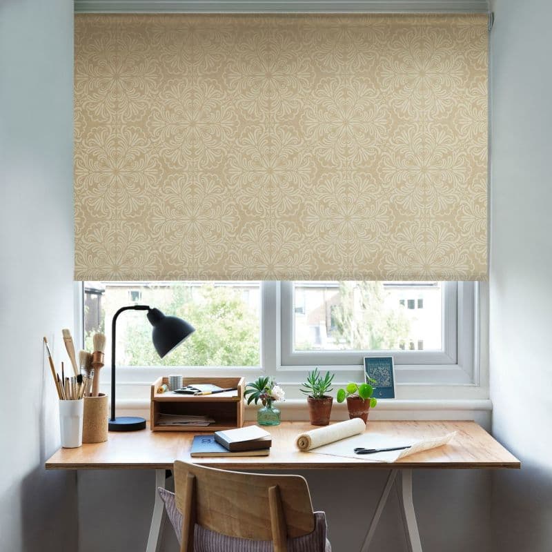 Beige patterned roller blind with ornate damask lowered over window, filtering daylight; above a wooden desk with a black lamp, paintbrushes, notebooks and small potted plants in a bright workspace.