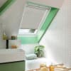 A white pleated skylight blind, mounted inside a sloped roof window, lowered to filter daylight; bathroom with mint-green trim, white herringbone tiles, sink, tub, plant and toiletries.