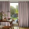 Floor-to-ceiling pleated striped curtains (gray and pink), parted centrally to reveal a glass door, filtering daylight into a dining room with wooden table, lamp, chair and sunlit garden beyond.