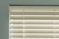 White horizontal slatted blinds occupying the top of a recessed window, partially closed with visible ladder tapes and pull cords, filtering bright daylight in a pale, softly lit room.