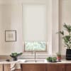 Light-colored roller blind lowered over a kitchen window, filtering daylight; stainless faucet and double sink below; countertop with bowls of leafy greens, potted plants, towel, and framed sketch on wall.