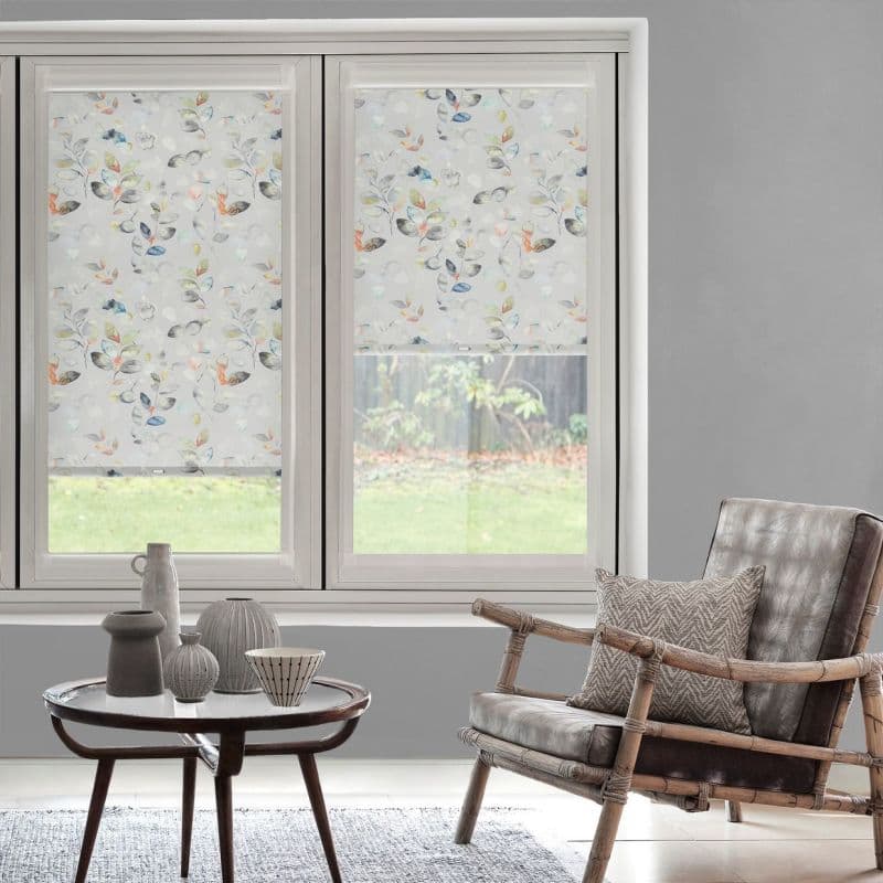 Patterned roller blinds with pastel watercolor leaf motif covering twin windows—left fully lowered, right partially raised—diffusing daylight into a grey living room with rattan leather armchair, table and backyard view.