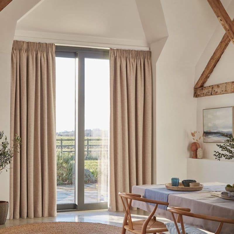 Beige full-length pleated curtains on a ceiling track, drawn to sides, framing glass patio doors and filtering soft daylight in a bright dining room with exposed wooden beams.