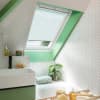 White pleated skylight blind, partially lowered across a slanted roof window, filtering soft daylight; bathroom with green-painted trim, white herringbone tiles, basin, bathtub ledge with plant and toiletries.