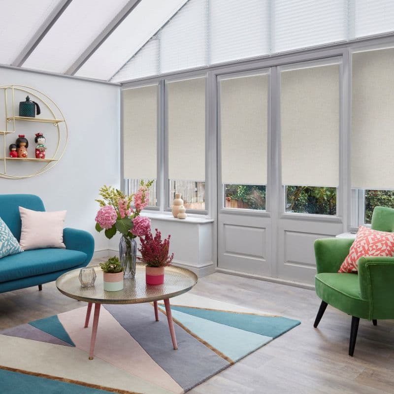 Beige roller blinds covering tall French windows and door, lowered to mid-height and filtering soft daylight; sunroom with glass roof, teal sofa, green armchair, and floral centerpiece on a coffee table.