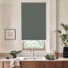 Dark gray roller blind covers most of a kitchen window above a stainless sink; wooden cabinets, countertop with a bowl of greens, potted plants and a framed sketch surround the window.