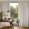 Floor-length beige pleated curtains drawn mostly closed with a central gap; filtering soft daylight and revealing a sunlit garden, foreground dining table set for breakfast.