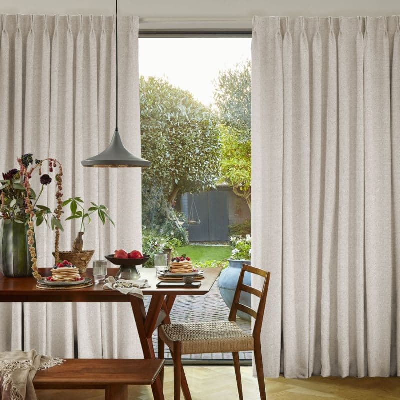 Floor-length beige pleated curtains drawn mostly closed with a central gap; filtering soft daylight and revealing a sunlit garden, foreground dining table set for breakfast.
