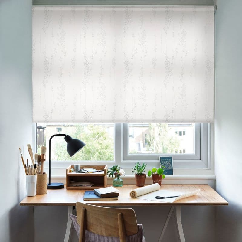 White roller blind with faint vertical floral pattern, lowered almost fully and filtering soft daylight over a wooden desk in a home study, with lamp, art brushes, notebooks, potted succulents.