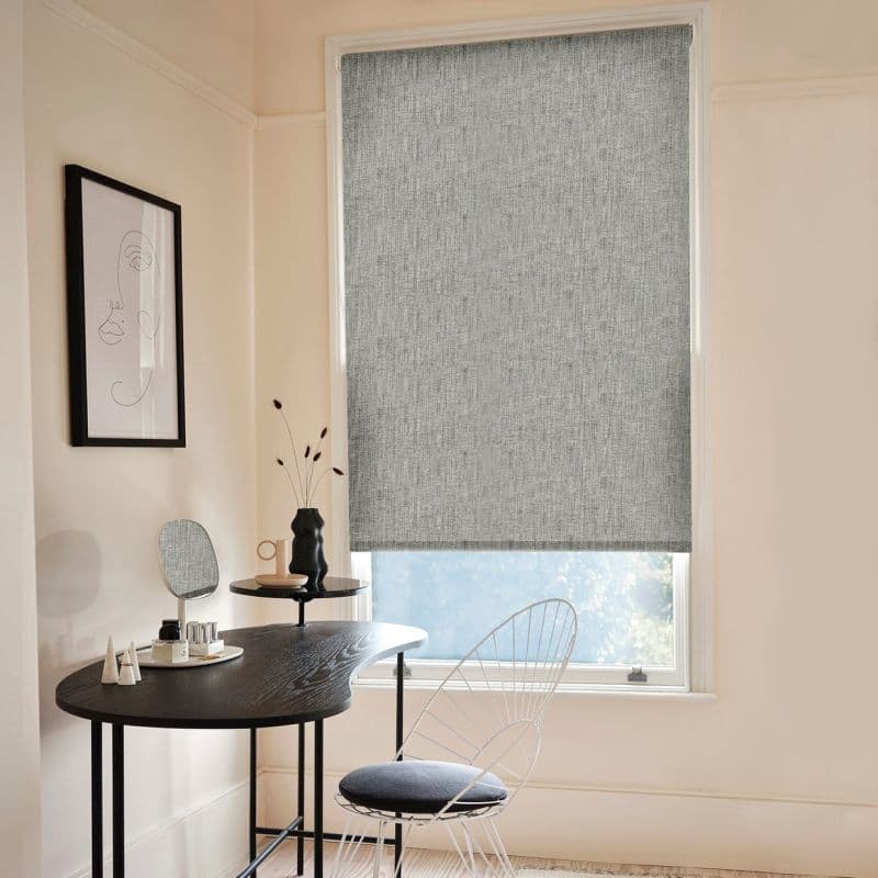 Textured grey roller blind lowered over a tall window, blocking and softly filtering daylight; neutral-toned dressing nook with black curvy vanity table, mirror, vase and wire chair.