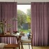 Thick mauve pleated floor-length curtains, parted to frame and filter soft daylight through the window; a dining table under a pendant lamp opens onto a sunlit garden with trees, shed.