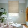 Beige patterned Roman blind, lowered over the window with a small bottom gap, filtering soft daylight; a pastel-green sitting nook with a white sculptural lounge chair, plant, floor lamp, and wooden floor.
