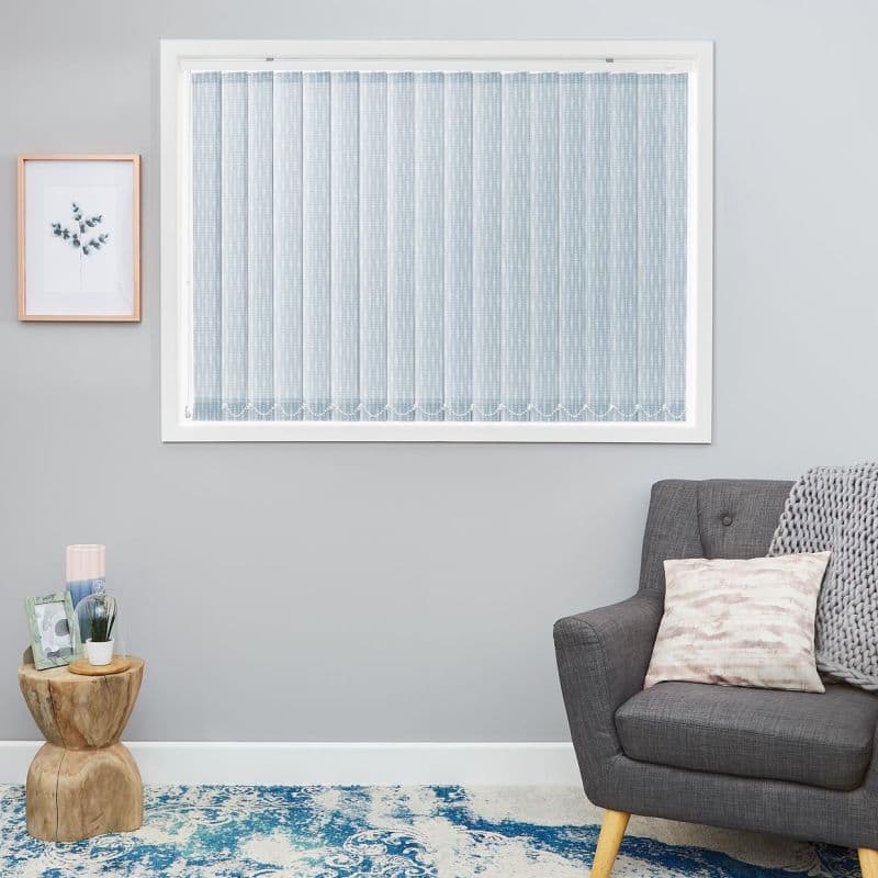 Sheer vertical blinds (pale blue, dotted) cover a rectangular window, fully closed and softly diffusing daylight; in a gray room with a charcoal armchair, wooden side table and blue rug.