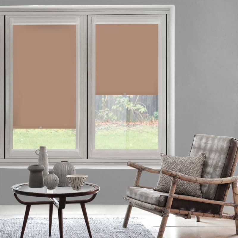 Two terracotta roller blinds cover twin windows—left fully lowered, right halfway—partially blocking and diffusing light in a neutral living room with a rattan armchair, coffee table, grassy backyard view.