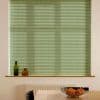 Pale green horizontal blinds, lowered and closed, diffusing daylight and obscuring the view; below them a sill with two bottles, a radiator, and a table with a bowl of oranges.