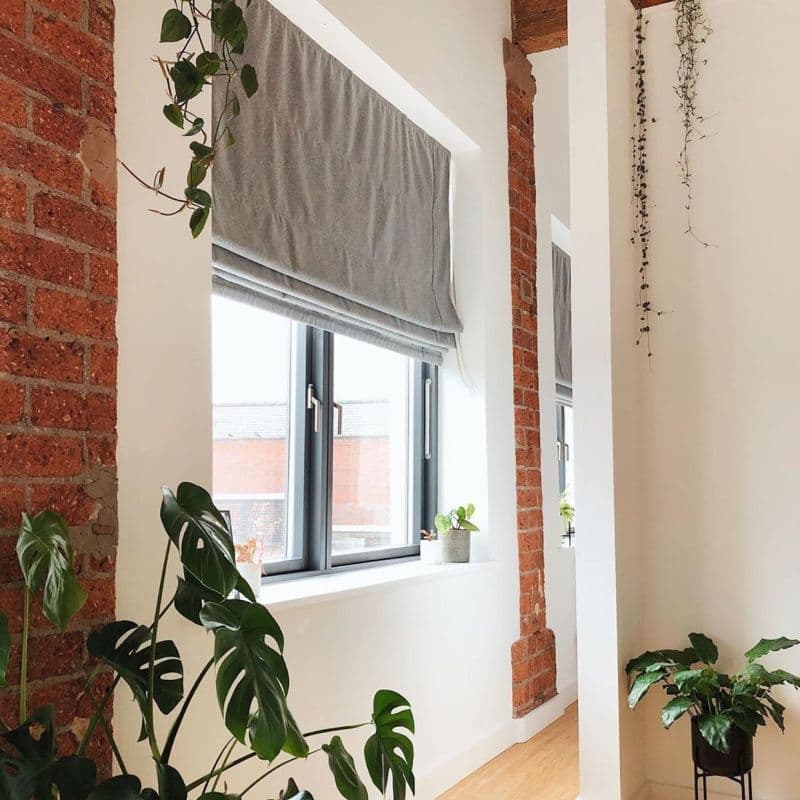 Gray Roman blind, partially lowered with soft folds, covering a black-framed window and filtering daylight. Bright minimalist living area with exposed red brick columns, wooden floor and potted plants.