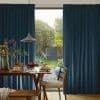 Teal pleated curtains, parted centrally, framing and filtering soft daylight; revealing a sunlit garden while a wooden dining table laid for breakfast and pendant lamp fill the cozy room.