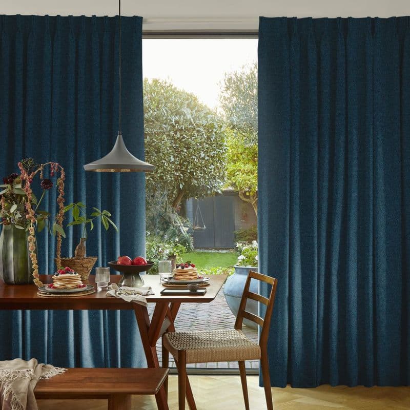 Teal pleated curtains, parted centrally, framing and filtering soft daylight; revealing a sunlit garden while a wooden dining table laid for breakfast and pendant lamp fill the cozy room.