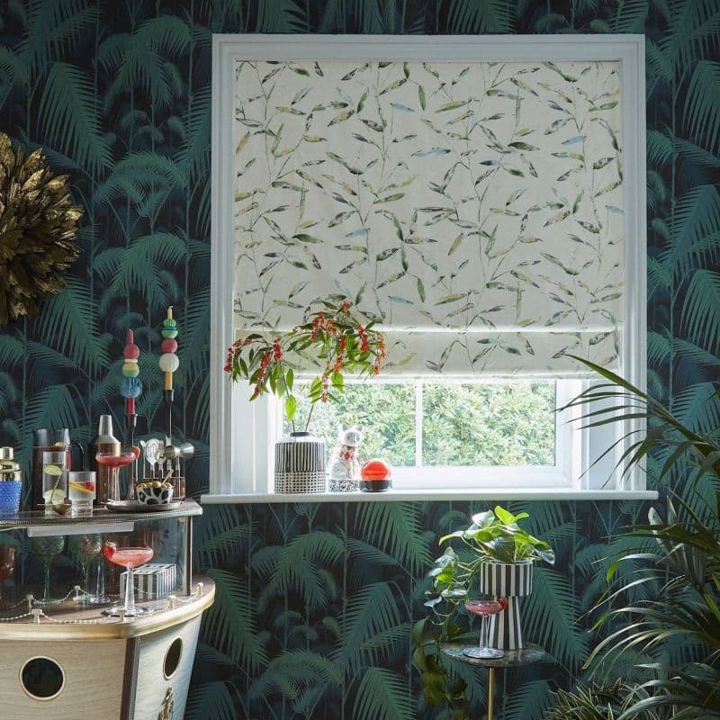 A leaf-printed Roman blind, mostly lowered across a white-framed window, filtering daylight; surrounded by dark palm wallpaper, houseplants, and a small bar cart.