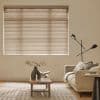 Beige horizontal slatted blinds covering a large window, tilted closed and softly filtering daylight; a beige sofa, wooden coffee table with sculptural vase and open book, black floor lamp, rug.