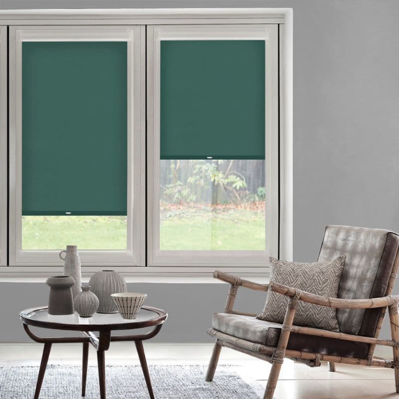 Teal roller blinds, two panels — left fully lowered, right partially lowered with pull tab — blocking light over window in a softly lit living room with a rattan armchair and table.
