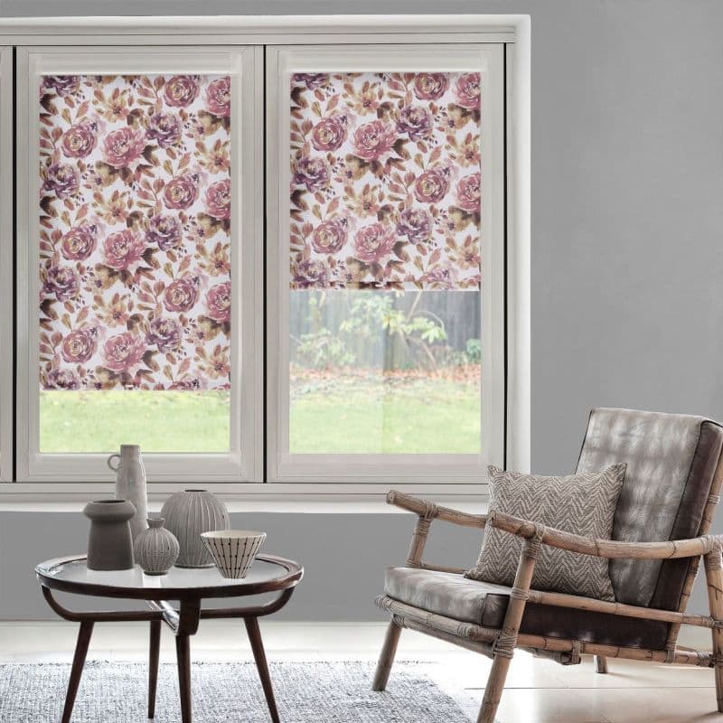 Floral roller blinds, lowered on a double window with pink-purple rose pattern, filtering daylight; set in a grey-walled living room with rattan armchair, round coffee table and garden view.