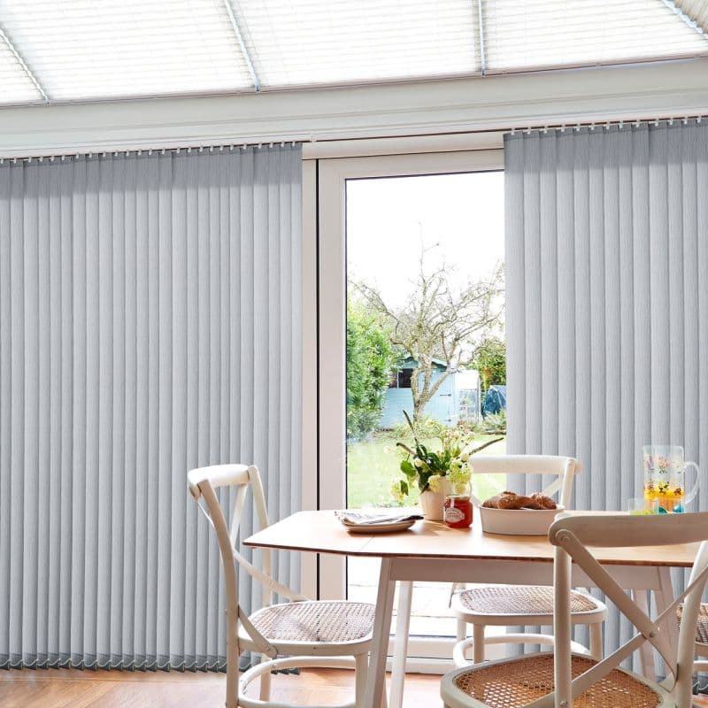 Pale grey vertical blinds with fabric slats, mostly closed across a glass door, parting centrally to admit light; sunlit dining nook with wooden table, cane chairs and flowers.