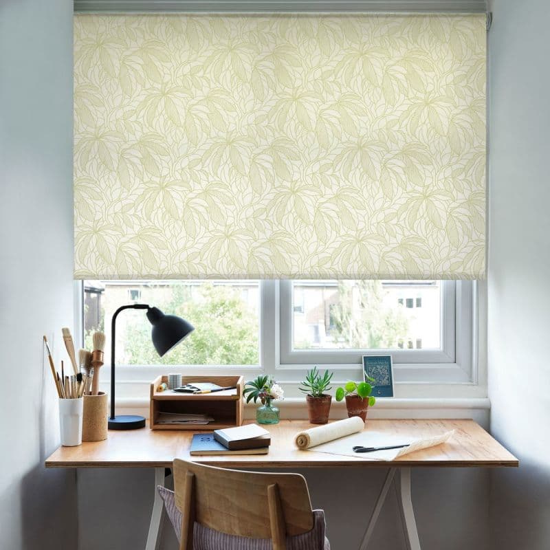 Patterned roller blind, pale cream with leaf print, lowered halfway; framing a bright home desk with lamp, brushes, potted succulents, chair and a daylight-filtered street view.