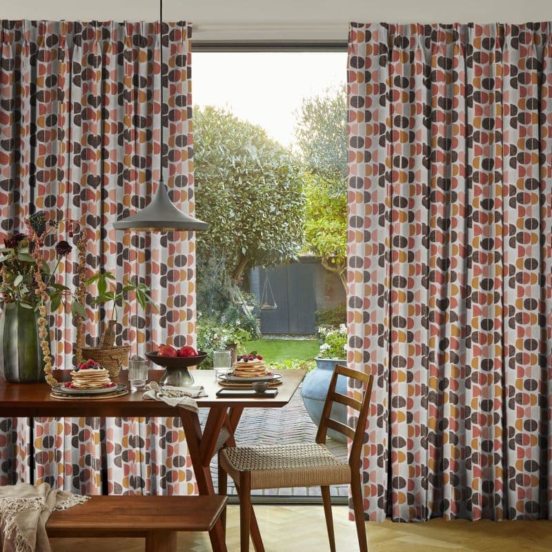 Full-length pleated patterned curtains (semi-circle design, orange, pink, brown) drawn to sides, framing a central opening that lets soft daylight through; dining table and pendant light face a green backyard.