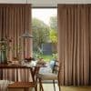 Floor-to-ceiling pleated brown curtains, drawn apart at center to frame a sunlit garden; filtering warm light over wooden dining table set with pancakes, pendant lamp and chair.