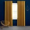 Mustard-gold grommet-top curtains, floor-length on a metal rod, are partially open, filtering bright daylight through a window against a deep navy wall with a gold wire chair and circular sconce.