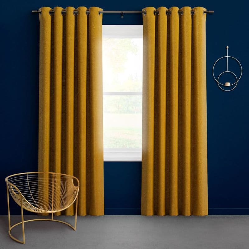 Mustard-gold grommet-top curtains, floor-length on a metal rod, are partially open, filtering bright daylight through a window against a deep navy wall with a gold wire chair and circular sconce.