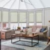 Light green pleated cellular blinds across tall angled conservatory windows, lowered to mid-height and softly filtering daylight; a bright seating area with sofa, wooden coffee table, rug, and plants.