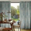 Full-length pleated blue-grey curtains, hanging on either side with soft folds — parted at the center, filtering morning light — framing a dining table set for breakfast and a sunlit garden.