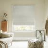 Light-gray textured Roman blind lowered with two soft folds, filtering diffuse daylight; in a neutral, cozy living room with sofa, wooden chest, knitted pouf and a leaning framed painting.