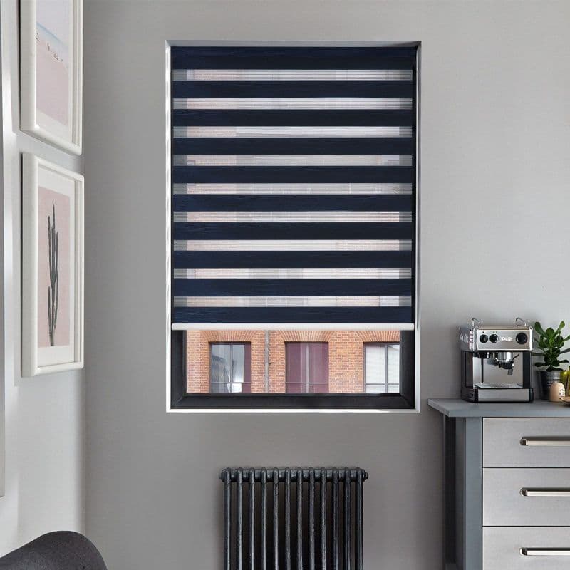Navy zebra roller blind lowered on window, alternating opaque and sheer bands filtering light with lower edge slightly raised; modern gray room corner with radiator below, espresso machine, brick view.