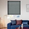Dark grey pleated blind, lowered over most of the window leaving a narrow bottom gap; living room with blue sofa, cushions, floor lamp, and roof tiles and trees visible beyond.