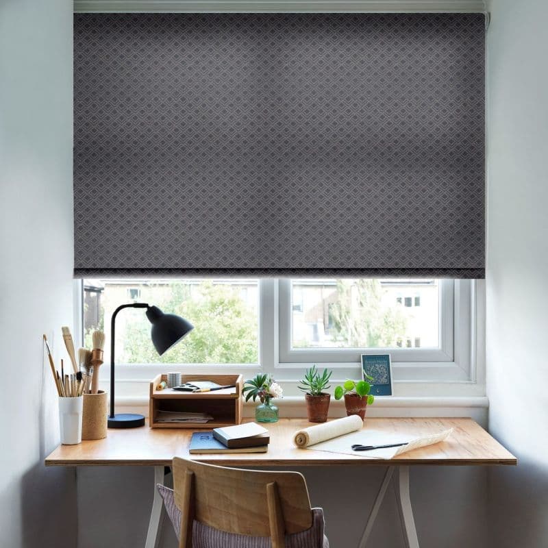 A patterned grey roller blind, fully lowered with a subtle diamond texture, covering a double window; blocking the view and softly diffusing daylight into a compact home desk area with lamp, plants, notebooks.