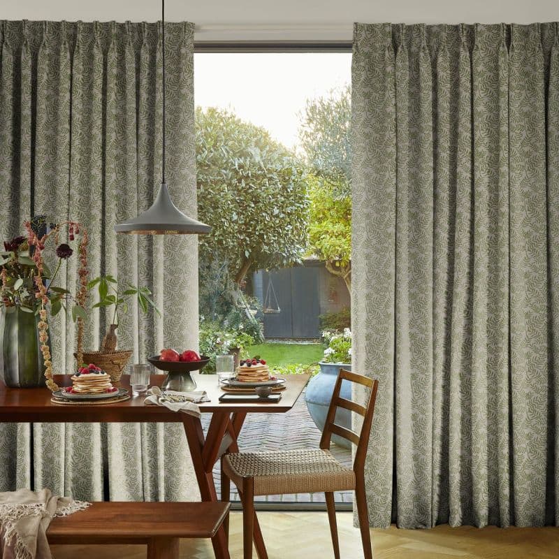 Full-length patterned curtains, beige with green fern print, parted to reveal sliding door; filtering soft daylight into a dining nook with wooden table set for breakfast and garden views.