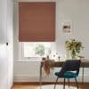 Terracotta Roman blind lowered over a sash window, folded at the bottom, filtering soft daylight into a home office with a wooden desk, teal chair, vase of greenery and walls.