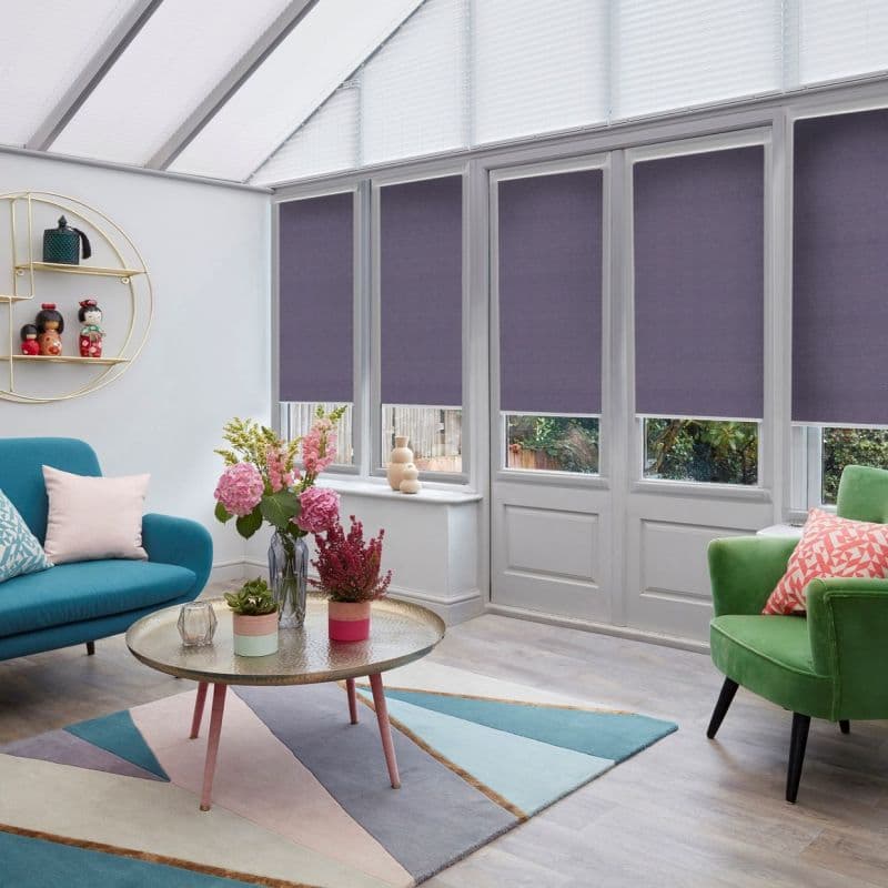 Plum roller blinds on tall windows and a door, lowered leaving narrow clear lower panes; filtering daylight in bright conservatory with pitched translucent roof, teal sofa, green armchair, pastel rug.