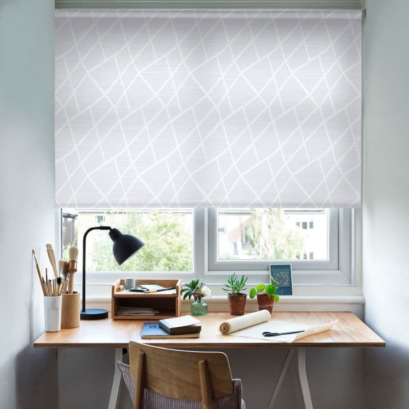 White patterned roller blind lowered across a double window, filtering daylight; a wooden desk below holds a black lamp, art brushes, potted succulents and stationery in a bright home workspace.