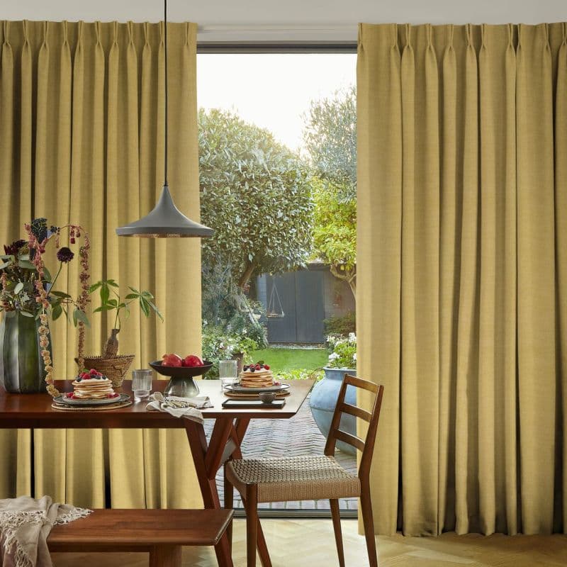 Floor-length pleated beige curtains, slightly parted at a sliding door, filtering daylight; wooden dining table set with pancakes and fruit beneath a pendant lamp, opening onto a lush backyard.