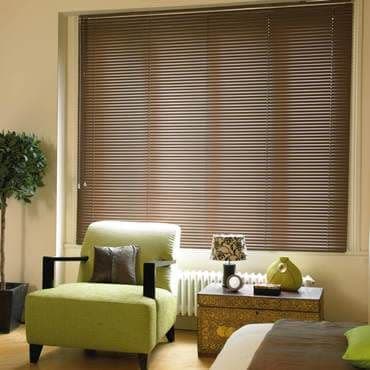 Brown horizontal slatted blinds cover a large window, fully closed filtering daylight; before them, lime-green armchair, patterned side table with lamp and potted tree—cozy living room.