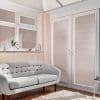 White horizontal Venetian blinds cover bay windows and French doors, closed and filtering sunlight into striped shadows; sunlit living room with pale tufted sofa, shelf, hardwood floor and cream rug.