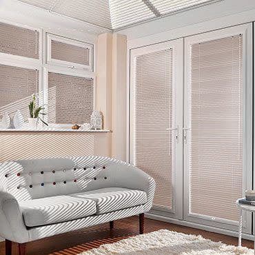 White horizontal Venetian blinds cover bay windows and French doors, closed and filtering sunlight into striped shadows; sunlit living room with pale tufted sofa, shelf, hardwood floor and cream rug.