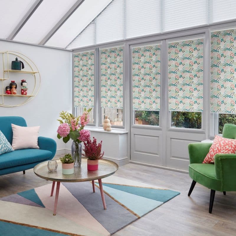 Floral-patterned roller blinds installed over tall conservatory windows and doors, lowered to filter daylight while leaving narrow lower gaps; bright sitting room with colorful sofas, round coffee table, and plants.
